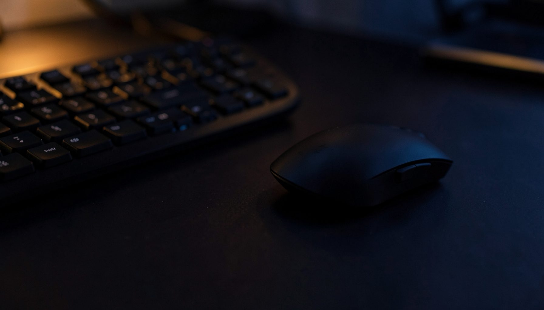 Dark desk surface with a wireless mouse resting beside a keyboard, subtle amber and navy ambient glow