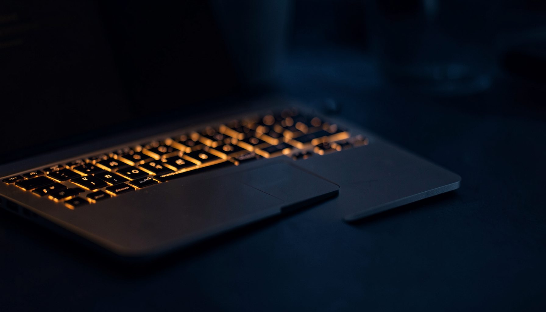 Dark workspace with a laptop keyboard glowing under warm amber light, deep navy and charcoal tones
