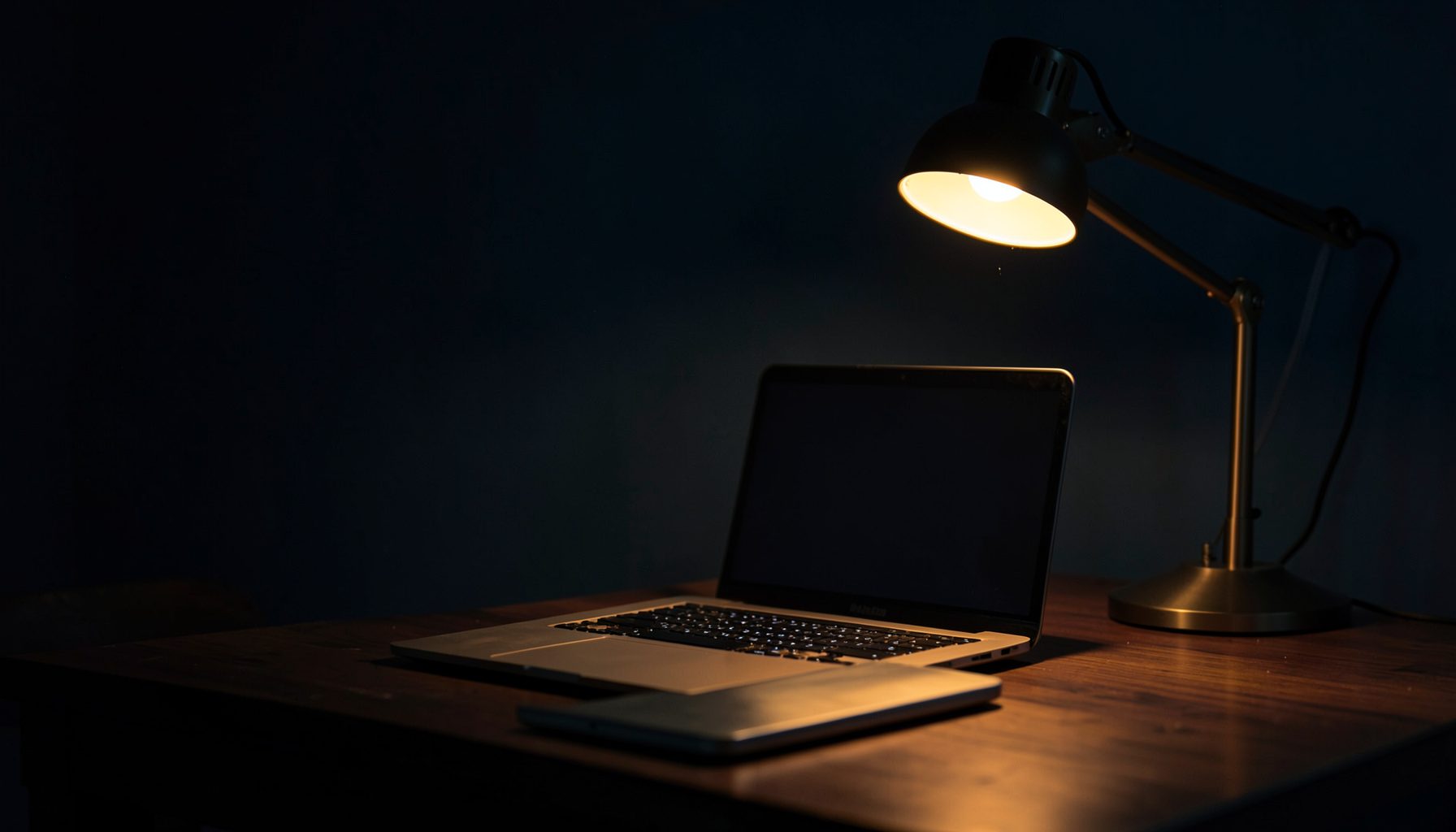 Dark workspace with a laptop on a wooden desk, warm amber desk lamp glow against deep navy shadows